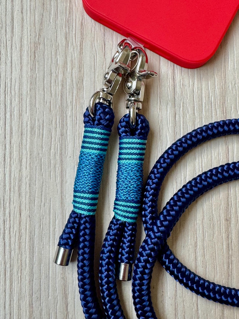 May include: Close-up of a navy blue phone lanyard with silver-tone metal clasps. The lanyard features a braided rope design with teal and blue striped accents. A red phone case is visible in the background.