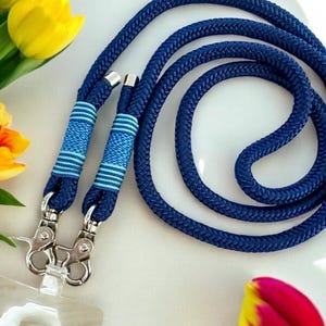 May include: A navy blue lanyard with silver-colored metal clasps. The lanyard is made of a thick, woven rope and features two sections with blue and white striped detailing. The lanyard is shown on a white surface with yellow tulips.