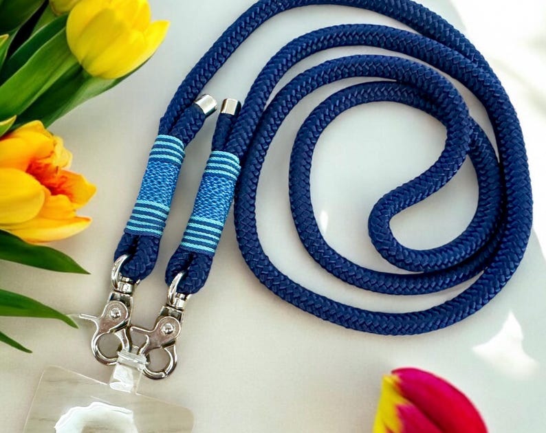 May include: A navy blue lanyard with silver-colored metal clasps. The lanyard is made of a thick, woven rope and features two sections with blue and white striped detailing. The lanyard is shown on a white surface with yellow tulips.