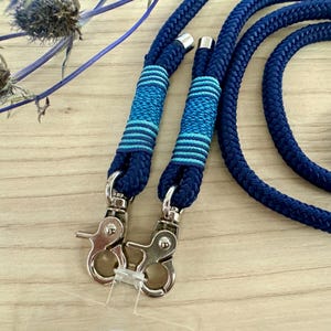 May include: Close-up of a navy blue rope leash with silver-colored metal clasps. The leash has decorative bands of blue and light blue stripes. The rope is thick and appears durable, suitable for pet accessories.