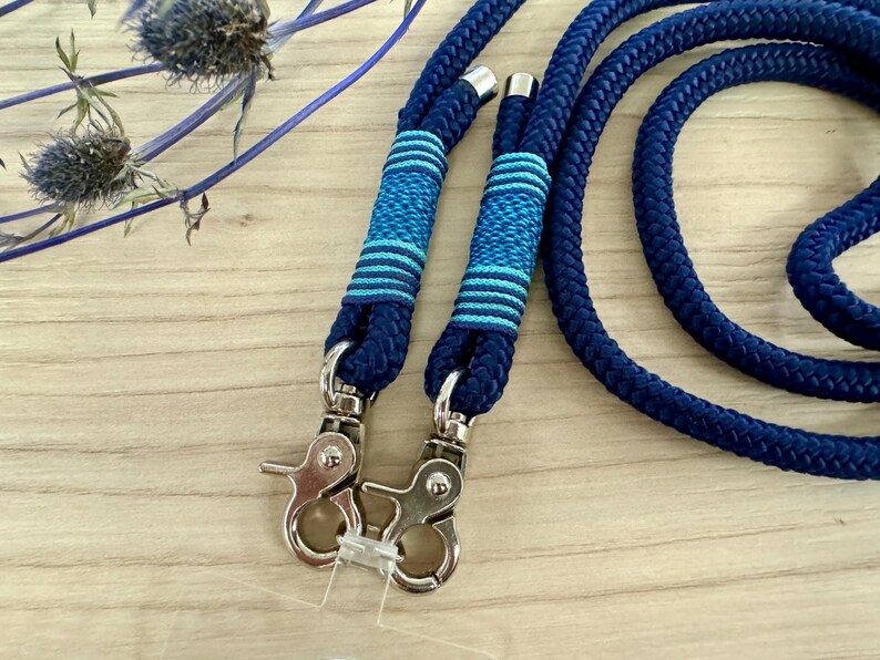 May include: Close-up of a navy blue rope leash with silver-colored metal clasps. The leash has decorative bands of blue and light blue stripes. The rope is thick and appears durable, suitable for pet accessories.