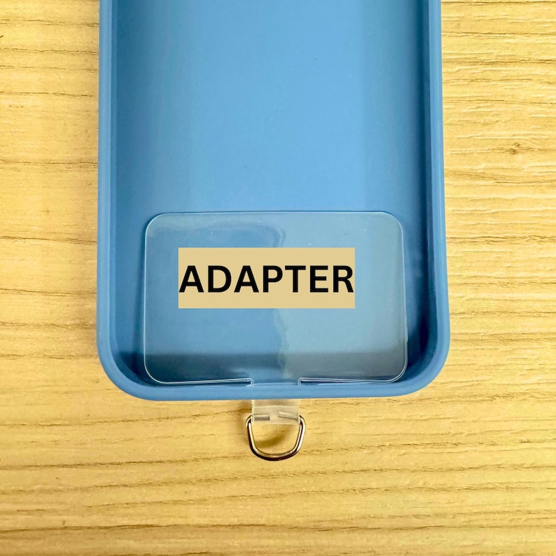 May include: A light blue phone case with a clear plastic adapter and a silver-colored metal ring. The word "ADAPTER" is printed on the adapter in black capital letters. The case is on a light brown wooden surface.