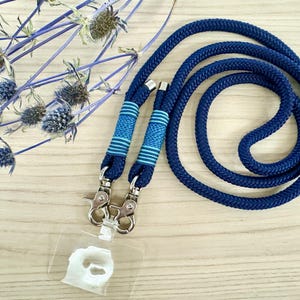 May include: A navy blue lanyard with a matching cord, featuring two decorative sections with blue and light blue stripes. The lanyard has silver-colored metal clasps and a clear plastic ID holder. Dried blue flowers are in the background.