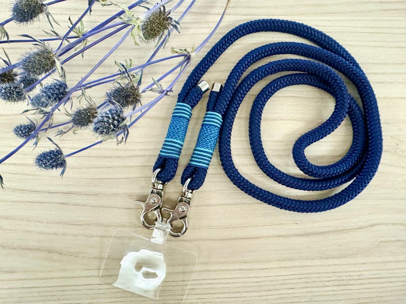May include: A navy blue lanyard with a matching cord, featuring two decorative sections with blue and light blue stripes. The lanyard has silver-colored metal clasps and a clear plastic ID holder. Dried blue flowers are in the background.