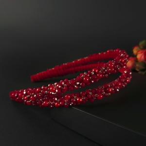 Könnte beinhalten: Ein rotes, perlenbesetztes Haarband mit Doppelband-Design. Das Haarband ist mit kleinen, runden und facettierten Perlen in verschiedenen Rottönen verziert. Die Perlen sind eng beieinander angeordnet und erzeugen einen strukturierten und schimmernden Effekt. Für besondere Anlässe.