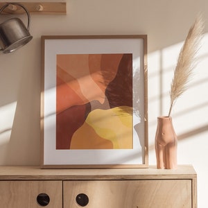 Może przedstawiać: Wzorowany wydruk w ramie w ciepłych kolorach, takich jak pomarańczowy, żółty i brązowy, wisi nad jasną drewnianą szafką z dwojgiem drzwi. Wysuszona łodyga trawy pampasowej znajduje się w jasnoróżowym ceramicznym wazonie.