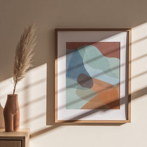 Puede incluir: Una impresión abstracta enmarcada con un diseño colorido de formas azules, verdes, naranjas y marrones. La impresión está colgada en una pared beige claro con una lámpara blanca y un jarrón con hierba de la pampa seca sobre una mesa de madera.
