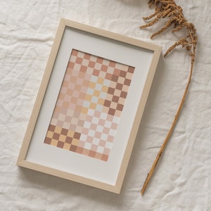 May include: A framed print of a geometric abstract design with a checkered pattern in shades of beige, brown, and pink. The print is in a light wood frame and is sitting on a white linen surface.