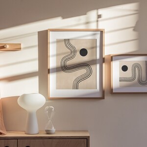 Puede incluir: Dos impresiones de arte abstracto enmarcadas con l&iacute;neas negras y c&iacute;rculos sobre un fondo beige. Las impresiones est&aacute;n colgadas en una pared beige claro con un perchero de madera y una l&aacute;mpara blanca sobre una mesa de madera debajo.