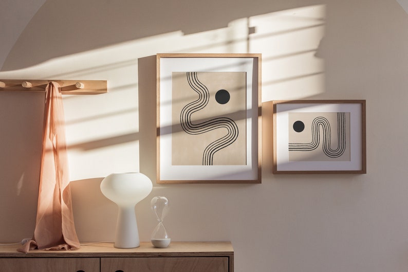 Puede incluir: Dos impresiones de arte abstracto enmarcadas con l&iacute;neas negras y c&iacute;rculos sobre un fondo beige. Las impresiones est&aacute;n colgadas en una pared beige claro con un perchero de madera y una l&aacute;mpara blanca sobre una mesa de madera debajo.