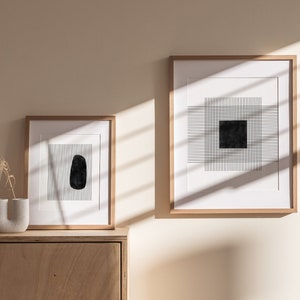 Puede incluir: Dos impresiones geométricas abstractas en blanco y negro enmarcadas con un marco de madera que cuelgan de una pared beige claro. Las impresiones son de estilo minimalista con formas y líneas geométricas.