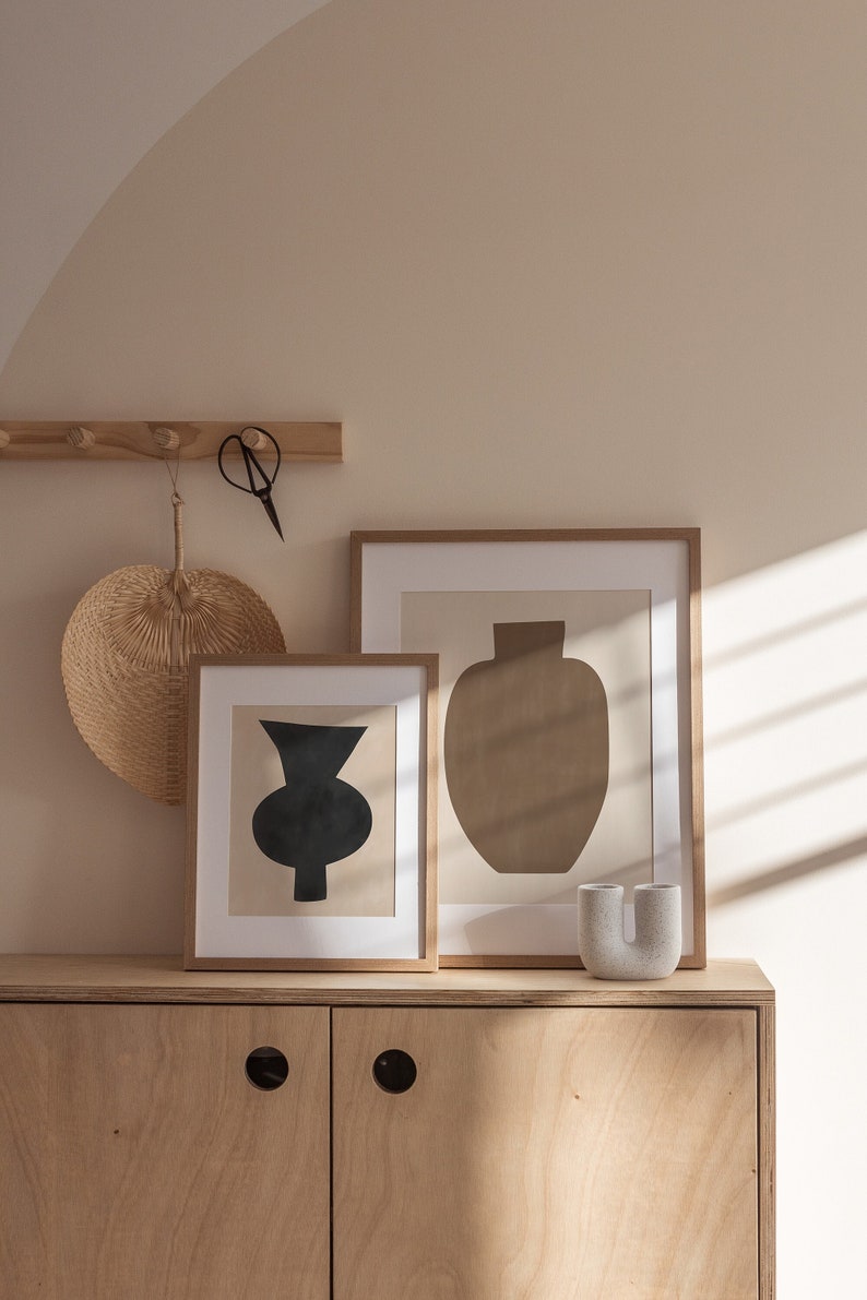 May include: Two framed abstract art prints with minimalist geometric shapes in black and brown, hanging on a beige wall above a light wood cabinet with two doors. A small, white ceramic vase sits on the cabinet.