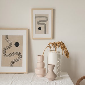 Puede incluir: Dos impresiones abstractas enmarcadas con l&iacute;neas negras y un c&iacute;rculo negro sobre un fondo beige. Tres jarrones, uno blanco, uno de vidrio transparente y uno rosa, est&aacute;n dispuestos sobre un mantel blanco con una rama de flores secas.