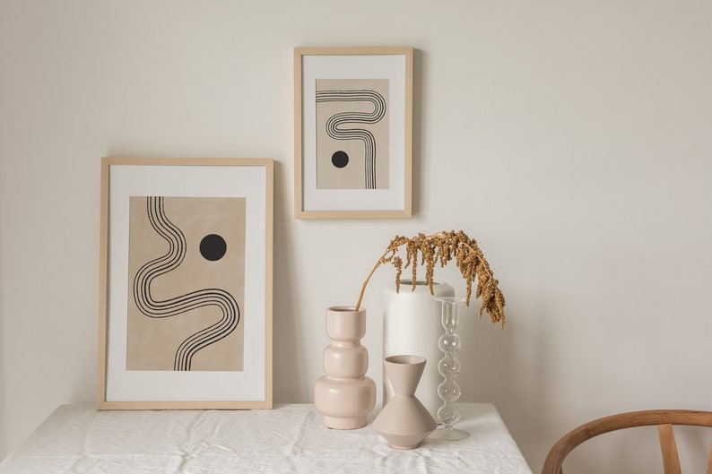 Puede incluir: Dos impresiones abstractas enmarcadas con l&iacute;neas negras y un c&iacute;rculo negro sobre un fondo beige. Tres jarrones, uno blanco, uno de vidrio transparente y uno rosa, est&aacute;n dispuestos sobre un mantel blanco con una rama de flores secas.