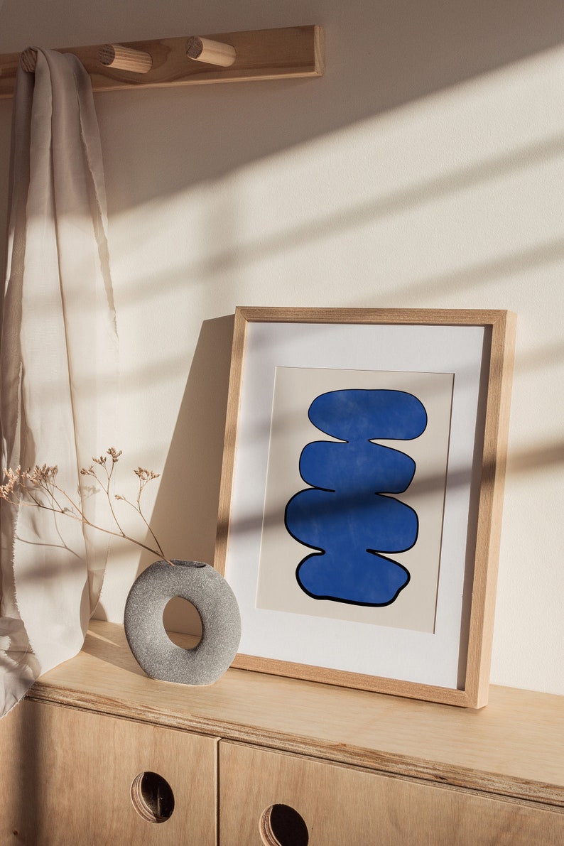 May include: A framed abstract print with a blue, organic shape design on a cream background. The print is displayed on a wooden shelf with a gray stone vase and dried flowers.