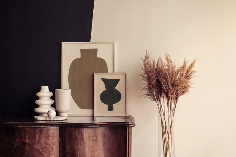 May include: Two framed abstract art prints of vases, one in brown and one in black, are displayed on a wooden dresser with a white ceramic vase and a small white ball. A tall glass vase with dried pampas grass is on the right side of the dresser.