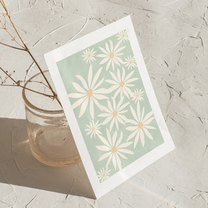 May include: A white card with a green background and a pattern of white daisies with yellow centers. The card is sitting on a white textured surface.