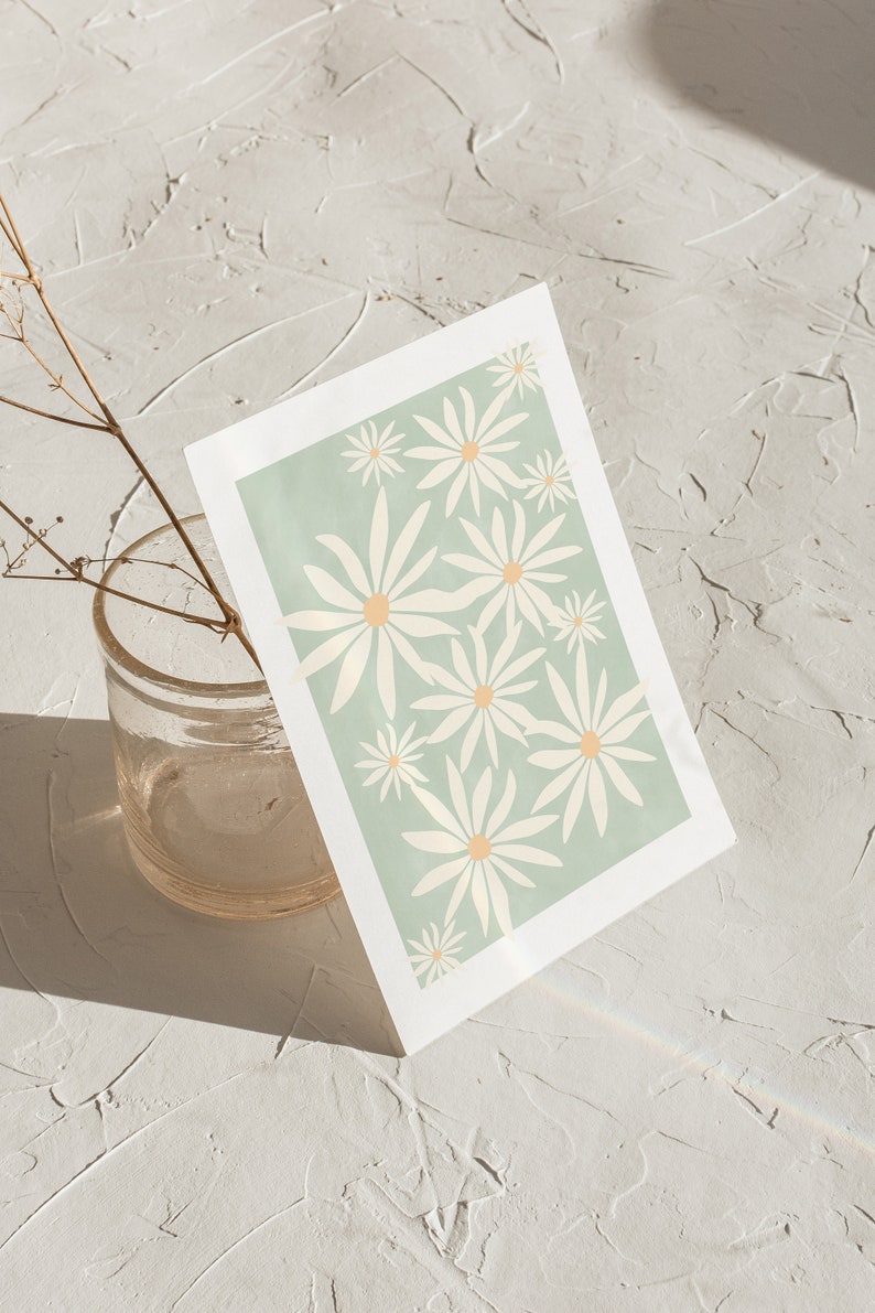 May include: A white card with a green background and a pattern of white daisies with yellow centers. The card is sitting on a white textured surface.