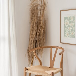 May include: A white framed print with a green background and white flowers. The print is hanging on a white wall next to a wooden chair with a woven seat. A dried grass plant is in the corner of the room.