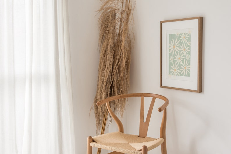 May include: A white framed print with a green background and white flowers. The print is hanging on a white wall next to a wooden chair with a woven seat. A dried grass plant is in the corner of the room.