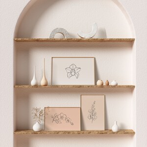 May include: Three minimalist line art prints of orchids in white frames on a wooden shelf with white and brown ceramic vases and decorative objects. The shelf is part of a white wall with an arched opening.