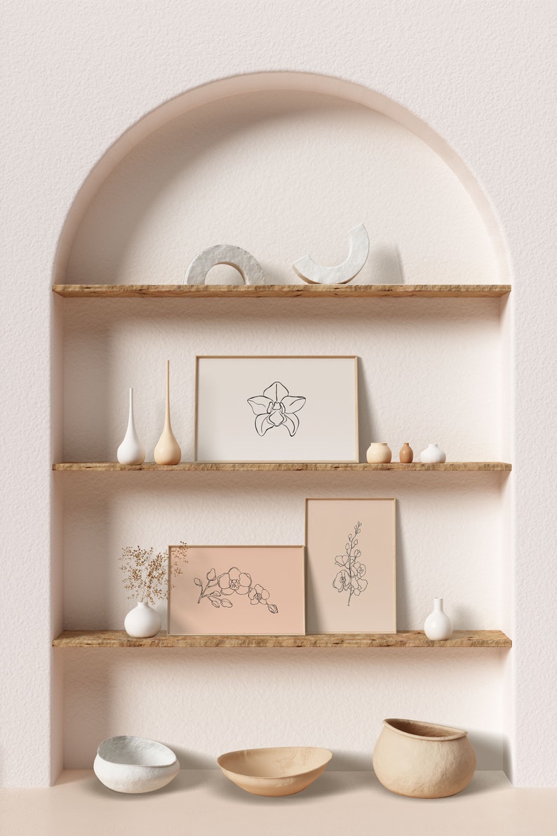 May include: Three minimalist line art prints of orchids in white frames on a wooden shelf with white and brown ceramic vases and decorative objects. The shelf is part of a white wall with an arched opening.