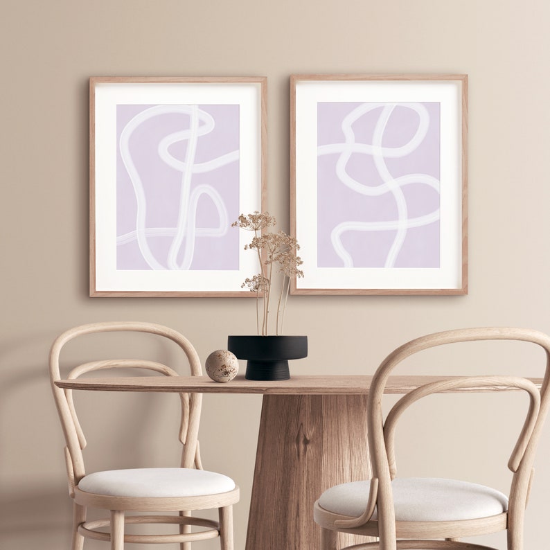 May include: Two framed abstract art prints with white lines on a light purple background. The prints are hanging on a light beige wall above a wooden table with two chairs.