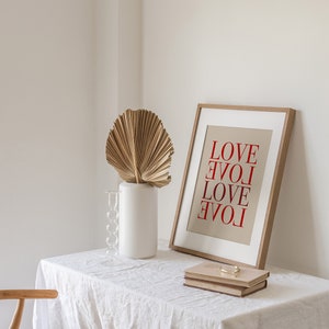 May include: A white table with a white linen tablecloth. On the table is a wooden frame with a beige background and the word "LOVE" written in red letters four times. There is a white vase with a dried palm leaf and a glass candle holder.