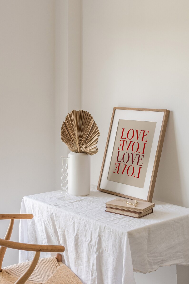 May include: A white table with a white linen tablecloth. On the table is a wooden frame with a beige background and the word "LOVE" written in red letters four times. There is a white vase with a dried palm leaf and a glass candle holder.
