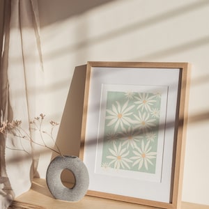 May include: A framed print with a light green background and a pattern of white daisies. The print is sitting on a wooden shelf with a gray stone vase and dried flowers.