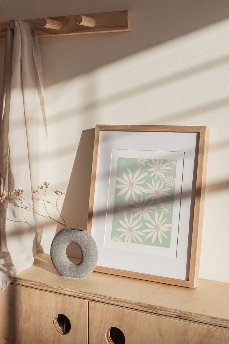 May include: A framed print with a light green background and a pattern of white daisies. The print is sitting on a wooden shelf with a gray stone vase and dried flowers.