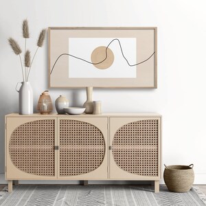 May include: A light wood cabinet with three doors and a woven rattan design. The cabinet is decorated with a vase of dried pampas grass, a lantern, and other decorative items. A framed print with a minimalist line drawing and a circle hangs above the cabinet.