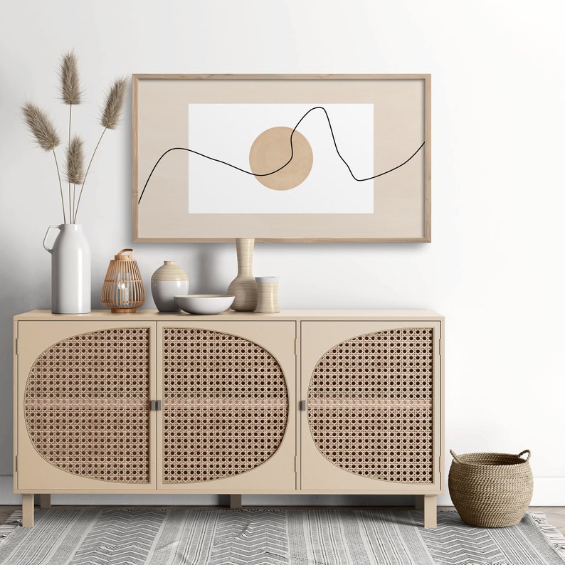 May include: A light wood cabinet with three doors and a woven rattan design. The cabinet is decorated with a vase of dried pampas grass, a lantern, and other decorative items. A framed print with a minimalist line drawing and a circle hangs above the cabinet.