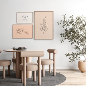 May include: Three framed line drawings of orchid flowers on a light pink background. The frames are light brown wood. The drawings are in black and white. The frames are hanging on a white wall. There is a table with four chairs in the foreground. The table and chairs are light brown. There is a potted plant in the background.
