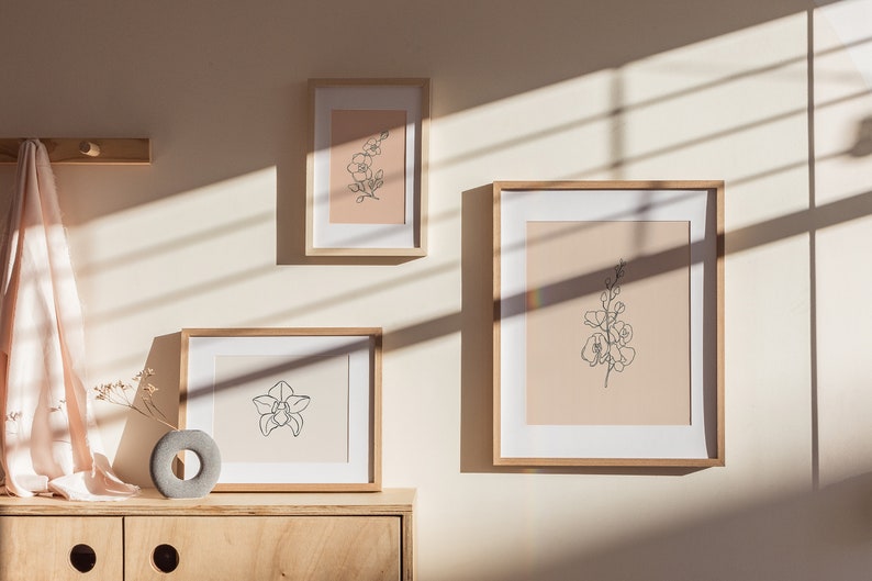 May include: Three framed prints of line drawings of flowers on a light beige wall. The prints are in wooden frames and are arranged in a staggered pattern. The wall is illuminated by sunlight streaming through a window, casting shadows on the wall.