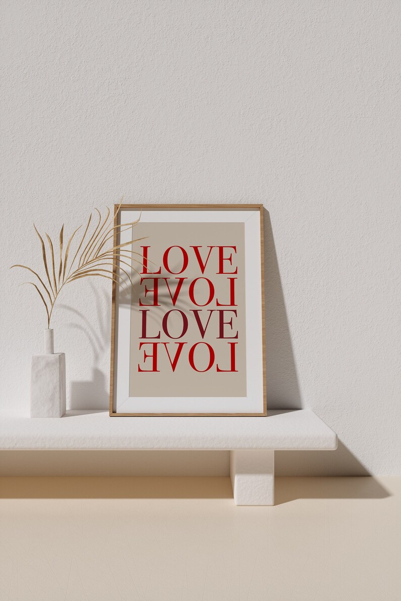 May include: A framed print with the word "LOVE" repeated four times, each time rotated 90 degrees. The print is on a beige background and is framed in a light wood frame. The print is sitting on a white shelf with a small vase with dried palm leaves.