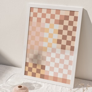 May include: A framed print with a checkered pattern in shades of brown, pink, and white. The print is sitting on a white tablecloth with a small pink vase and a dried flower stem.