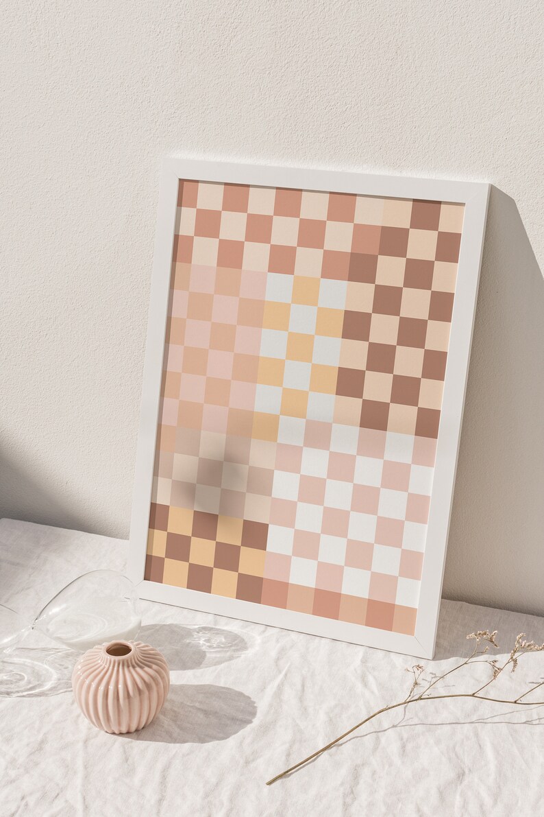 May include: A framed print with a checkered pattern in shades of brown, pink, and white. The print is sitting on a white tablecloth with a small pink vase and a dried flower stem.