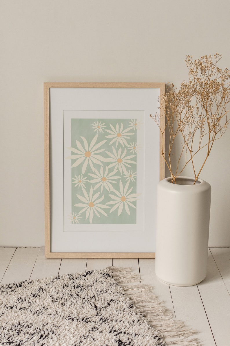 May include: A framed print of a floral design with white flowers on a light green background. The frame is made of light wood and is sitting on a white floor with a fluffy rug in front of it.