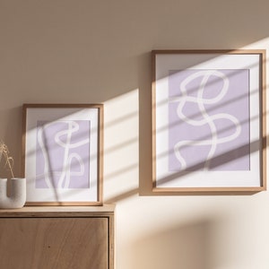 May include: Two framed abstract art prints with a light purple background and white line drawings. The prints are hanging on a light beige wall with a wooden shelf above them.