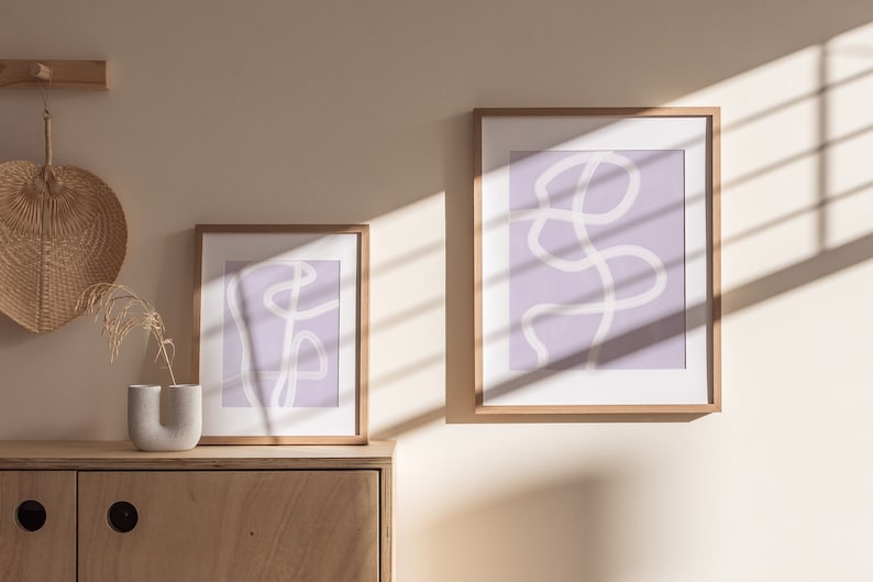 May include: Two framed abstract art prints with a light purple background and white line drawings. The prints are hanging on a light beige wall with a wooden shelf above them.