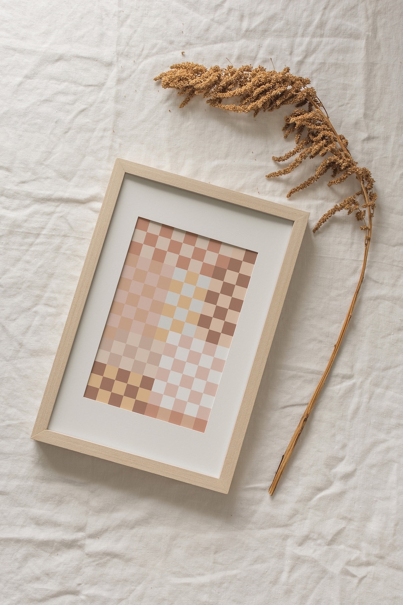 May include: A framed print of a geometric abstract design with a checkered pattern in shades of beige, brown, and pink. The print is in a light wood frame and is sitting on a white linen surface.