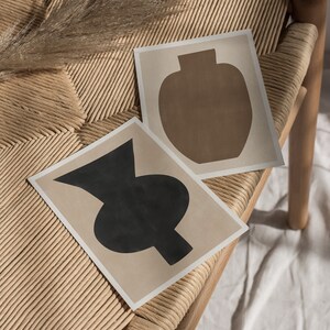 May include: Two abstract art prints of vases on a light brown wicker chair. The vase on the left is black and the vase on the right is brown.