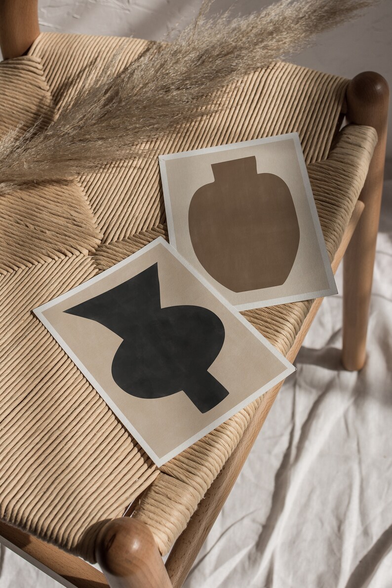 May include: Two abstract art prints of vases on a light brown wicker chair. The vase on the left is black and the vase on the right is brown.