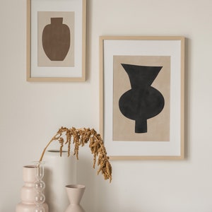 May include: Two framed abstract art prints with minimalist vase designs in black and brown on a beige background. The prints are hanging on a white wall above a table with three ceramic vases and a dried flower arrangement.