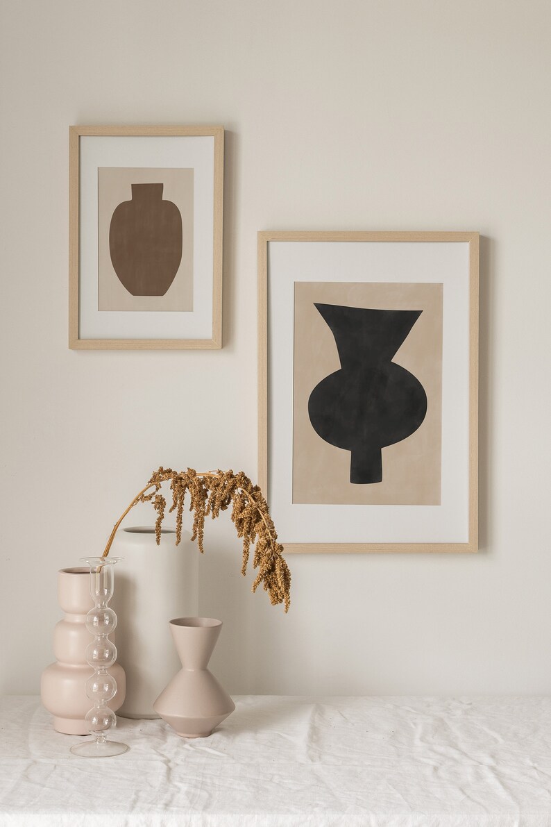 May include: Two framed abstract art prints with minimalist vase designs in black and brown on a beige background. The prints are hanging on a white wall above a table with three ceramic vases and a dried flower arrangement.