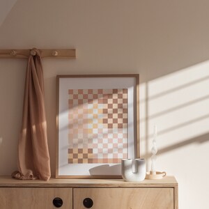 May include: A light wood cabinet with two doors and a light brown scarf hanging on a wooden rack above. A framed print with a checkered pattern in shades of pink, yellow, and brown is on top of the cabinet. A white ceramic vase and a white candle are also on top of the cabinet.