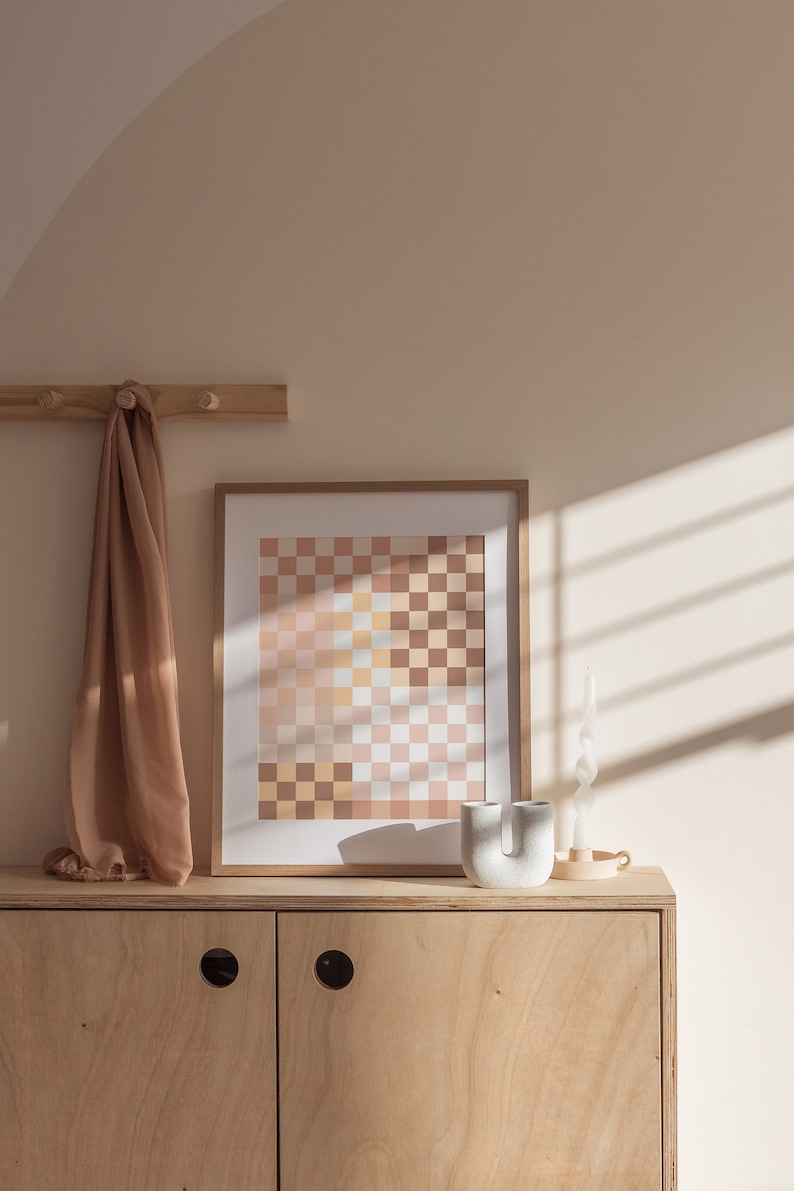 May include: A light wood cabinet with two doors and a light brown scarf hanging on a wooden rack above. A framed print with a checkered pattern in shades of pink, yellow, and brown is on top of the cabinet. A white ceramic vase and a white candle are also on top of the cabinet.