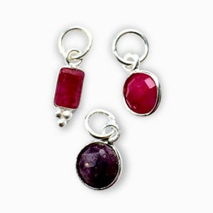 May include: Three silver charms with red gemstones. One charm has a rectangular ruby, another has an oval ruby, and the third has a round, dark red gemstone. Each charm has a small silver loop for attachment.