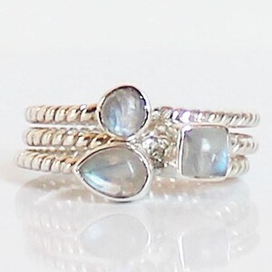 May include: A stack of three silver rings featuring twisted bands. Each ring is topped with a single moonstone in a different shape: one round, one pear-shaped, and one square. The stones exhibit a translucent, iridescent quality with blue flashes.